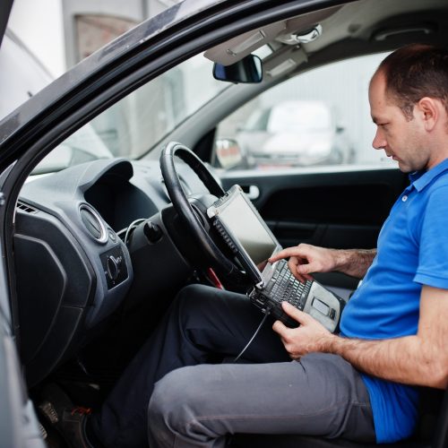 Car repair and maintenance theme. Electric mechanic in uniform working in auto service, making car diagnostics using obd device with laptop.